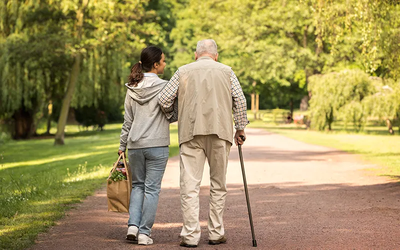 A senior citizen with a can being assisted by a younger woman on a walk through a park.